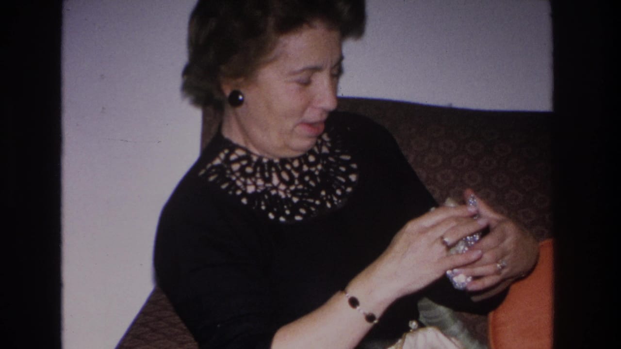 Middle-aged woman in black dress with cutwork neckline handling a small glittering object, seated indoors.
