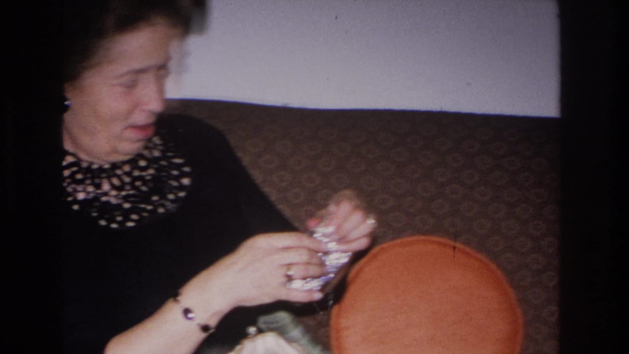 Woman in black dress with decorative neckline continues working a small object in her hands; orange cushion prominent at right; Hoboken, New Jersey, 1966 8mm home movie.