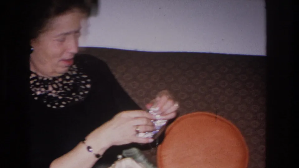 Woman in black dress with decorative neckline continues working a small object in her hands; orange cushion prominent at right; Hoboken, New Jersey, 1966 8mm home movie.