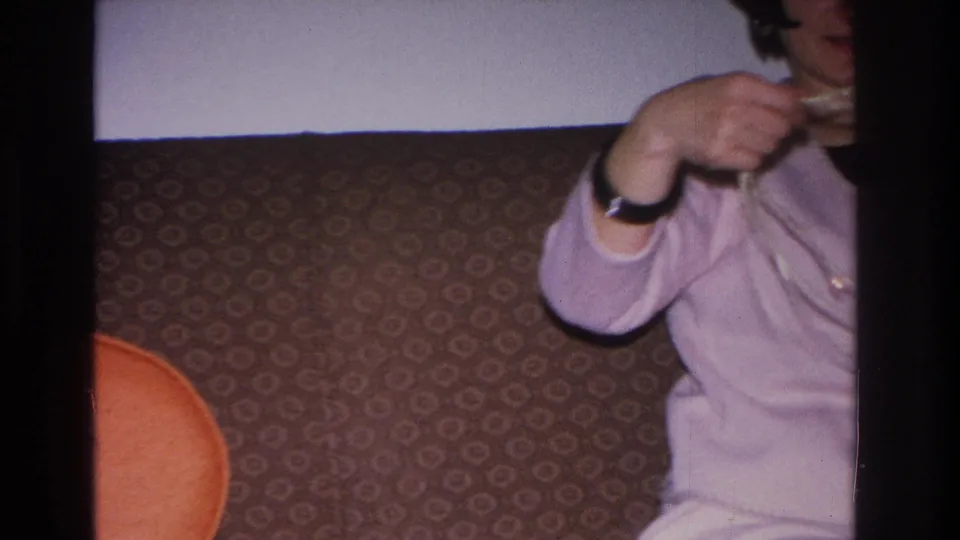 Woman in light pink sweater handles a long strand of yarn or beads during a domestic craft session; Hoboken, New Jersey, 1966 8mm home movie.