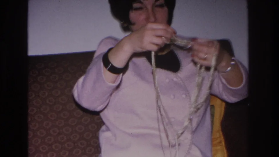Woman in light pink sweater threads or works a long bead or yarn strand while seated in a yellow-cushioned chair; Hoboken, New Jersey, 1966 8mm home movie.