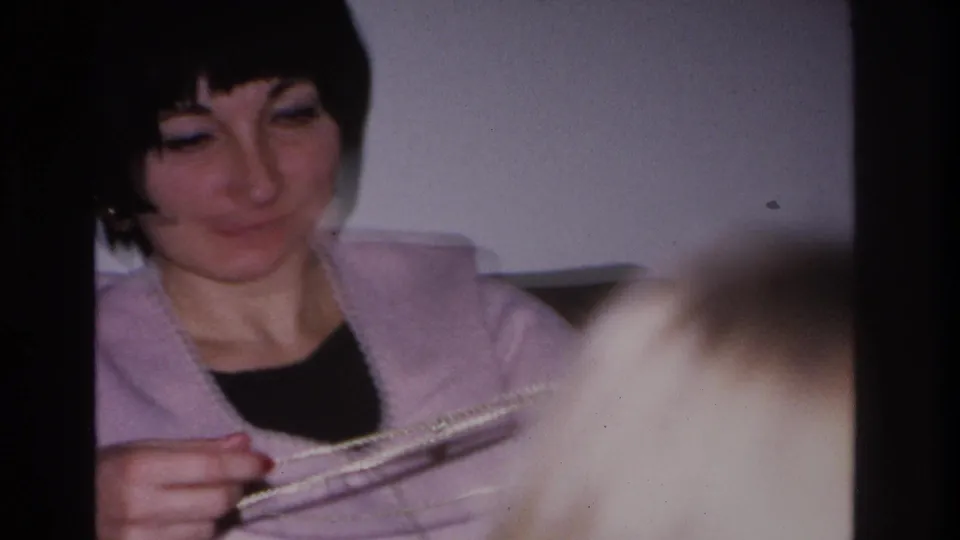 Woman in lavender cardigan examines beaded string held low in frame; unidentified blurred object enters foreground; 8mm home movie, 1966.