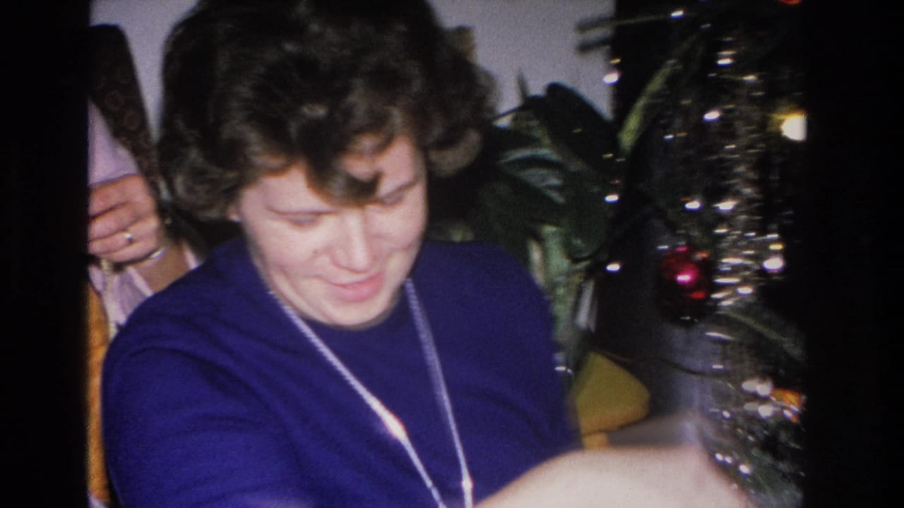Woman in a blue sweater and pendant necklace looks downward with a gentle expression; a tinsel Christmas tree with red ornaments is visible behind her at right.