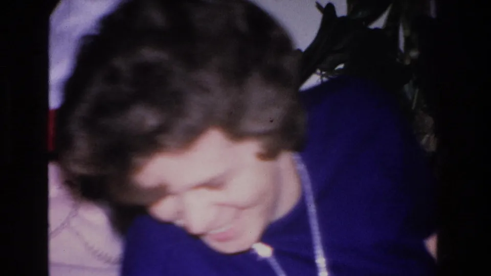 Tight close-up of a woman with dark curly hair and pendant necklace looking downward at a 1966 holiday gathering, with Christmas tree tinsel partially visible at the top right.