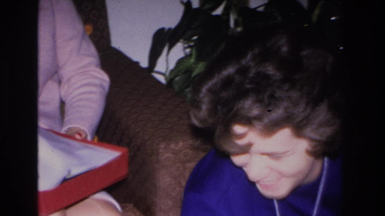 Woman in blue top and long pendant necklace looks down smiling at an open red gift box, 8mm home movie circa 1966.