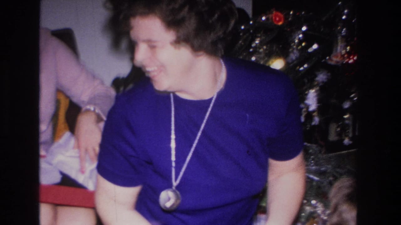 Woman in blue top and pendant necklace smiling toward someone off-camera, Christmas tree with tinsel at right.