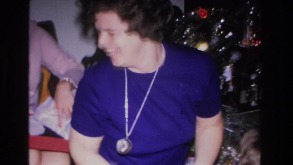 Woman in blue top smiling off-camera beside a tinsel-decorated Christmas tree with colored ornaments, 8mm home movie circa 1966.