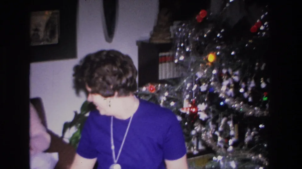 Woman in blue top viewed from side as camera pans to reveal heavily tinsel-draped Christmas tree, bookshelf visible in background, 8mm home movie circa 1966.