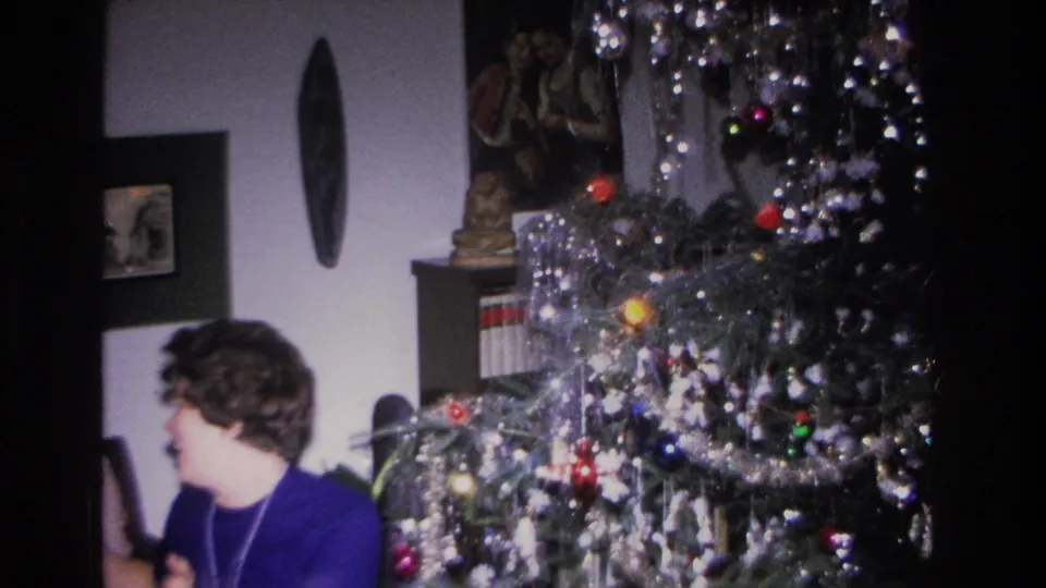 Woman in blue top looking toward camera beside a heavily tinsel-decorated Christmas tree, bookshelf, figurine, and circular wall decoration visible, 8mm home movie circa 1966.
