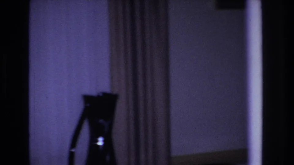 Opening 8mm frame from a 1966 Hoboken home movie: a dim living room with a pronounced purple dye-cast, a silhouetted tripod-mounted object, and floor-length drawn curtains.