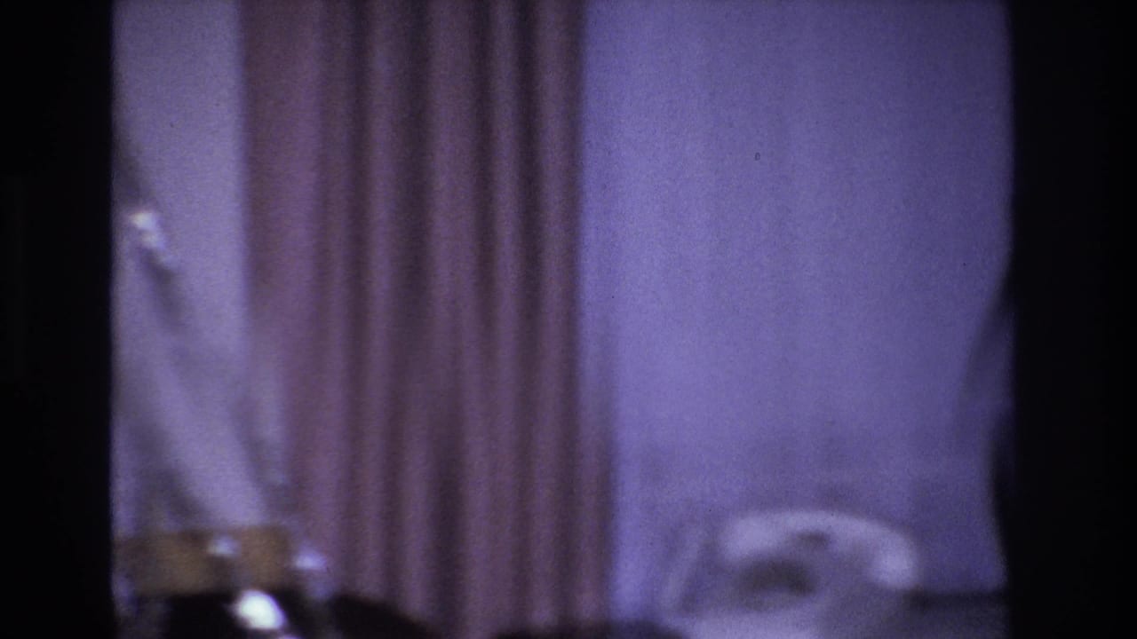Purple-hued room interior mid-pan showing curtains, unidentified dark objects at the lower frame edge, and a partial white chair at right.