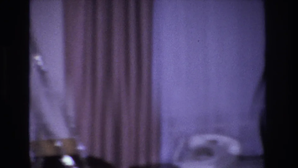 Mid-pan 8mm frame from a 1966 Hoboken home movie: purple-cast room with drawn curtains, ambiguous dark cylindrical objects at lower frame, and a partial white chair visible at right.