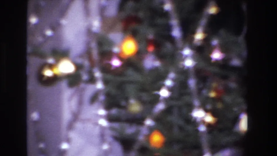 Final 8mm frame of the 1966 Christmas tree: heavy bokeh with a dominant warm orange-yellow incandescent bulb at center, red ornaments, and tinsel strands against a purple-cast interior wall.