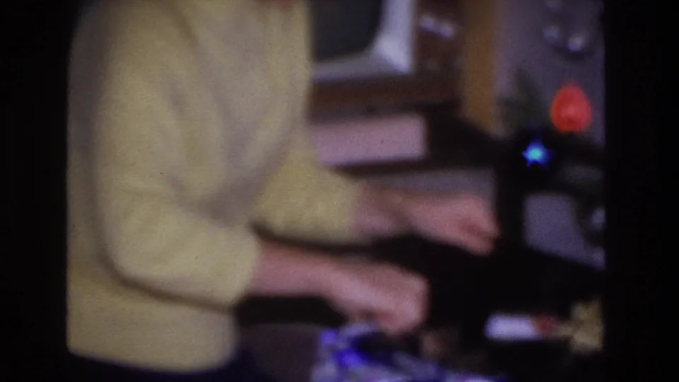 Close view of woman in pale yellow sweater handling a dark-wrapped gift, blue and red ornaments visible at right edge, circa 1966 8mm home movie