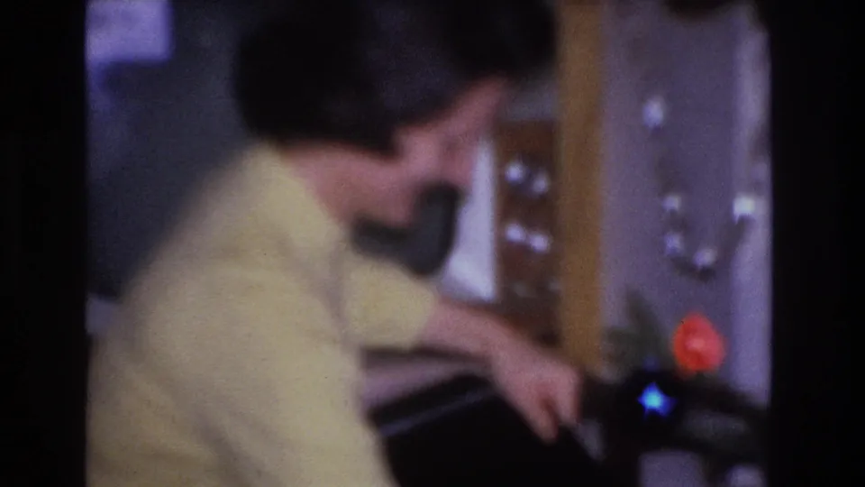 Woman in pale yellow knit sweater bending forward to open a dark gift box, Christmas tree with red and blue ornament lights behind her, circa 1966 8mm
