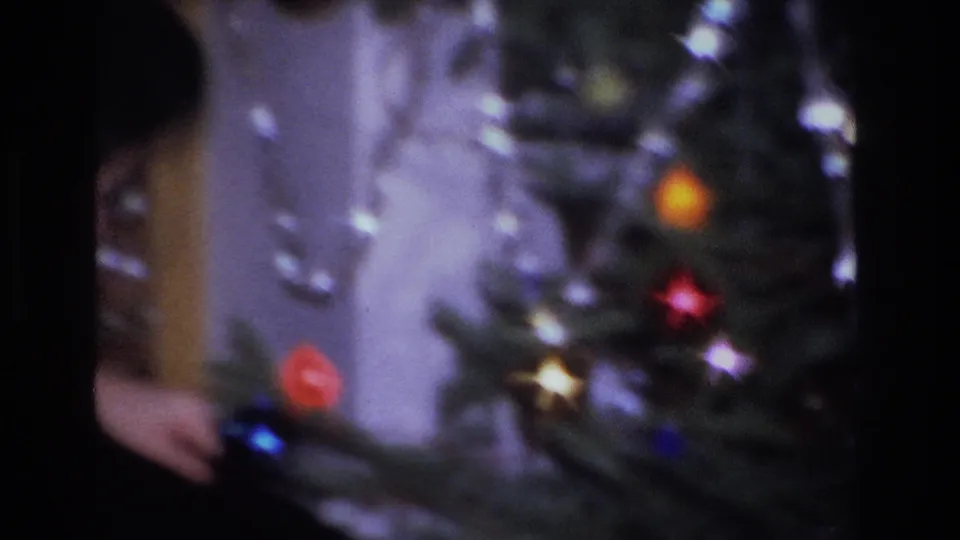 Close view of decorated Christmas tree with orange, red, and white incandescent bulbs; woman's arms in pale yellow sweater at lower left, circa 1966 8mm