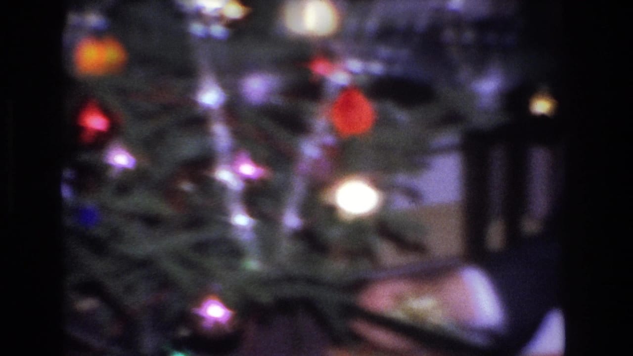 Man in a dark suit bent toward the base of a decorated Christmas tree; colored globe ornaments and incandescent string lights visible; Hoboken, New Jersey home, circa 1966.