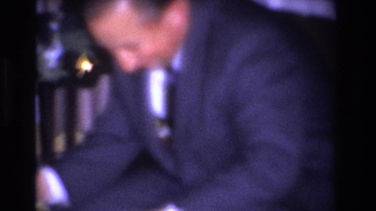Close soft-focus shot of a man in a dark suit and tie leaning forward; dark stair balusters visible in background.