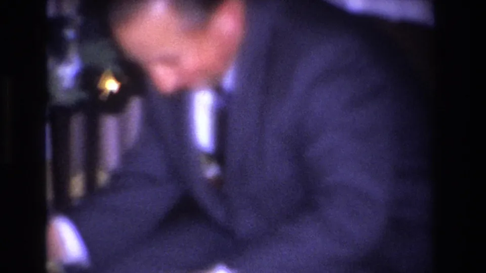 Blurred close-up of a man in a dark suit and white dress shirt leaning forward; stair balusters and warm light visible in background; 1960s home interior, circa 1966.