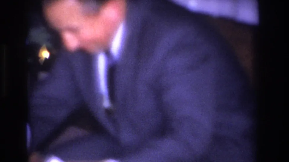 Extreme close-up of a man in a dark jacket, heavily out of focus; a glowing Christmas tree light at lower left edge; 8mm home movie, circa 1966.