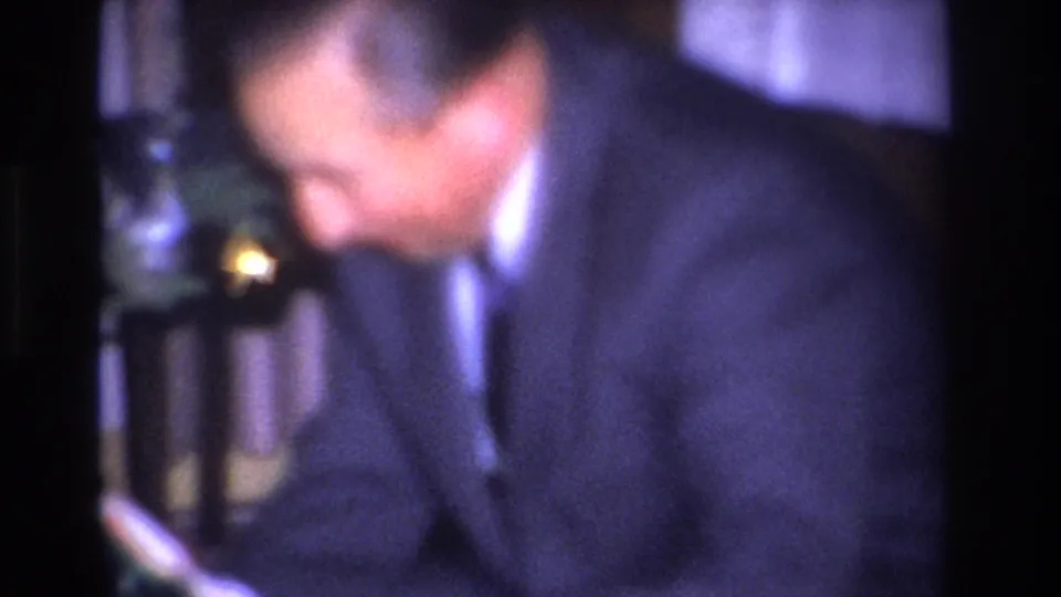 Man in dark suit and white shirt looking down at an object in his hands; Christmas tree with colored ornaments at right; Hoboken, New Jersey home interior, circa 1966.