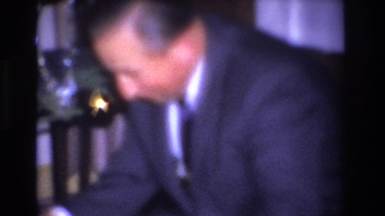Close-up of a formally dressed man in a dark suit and tie looking downward near a decorated Christmas tree, from a 1960s 8mm home movie.