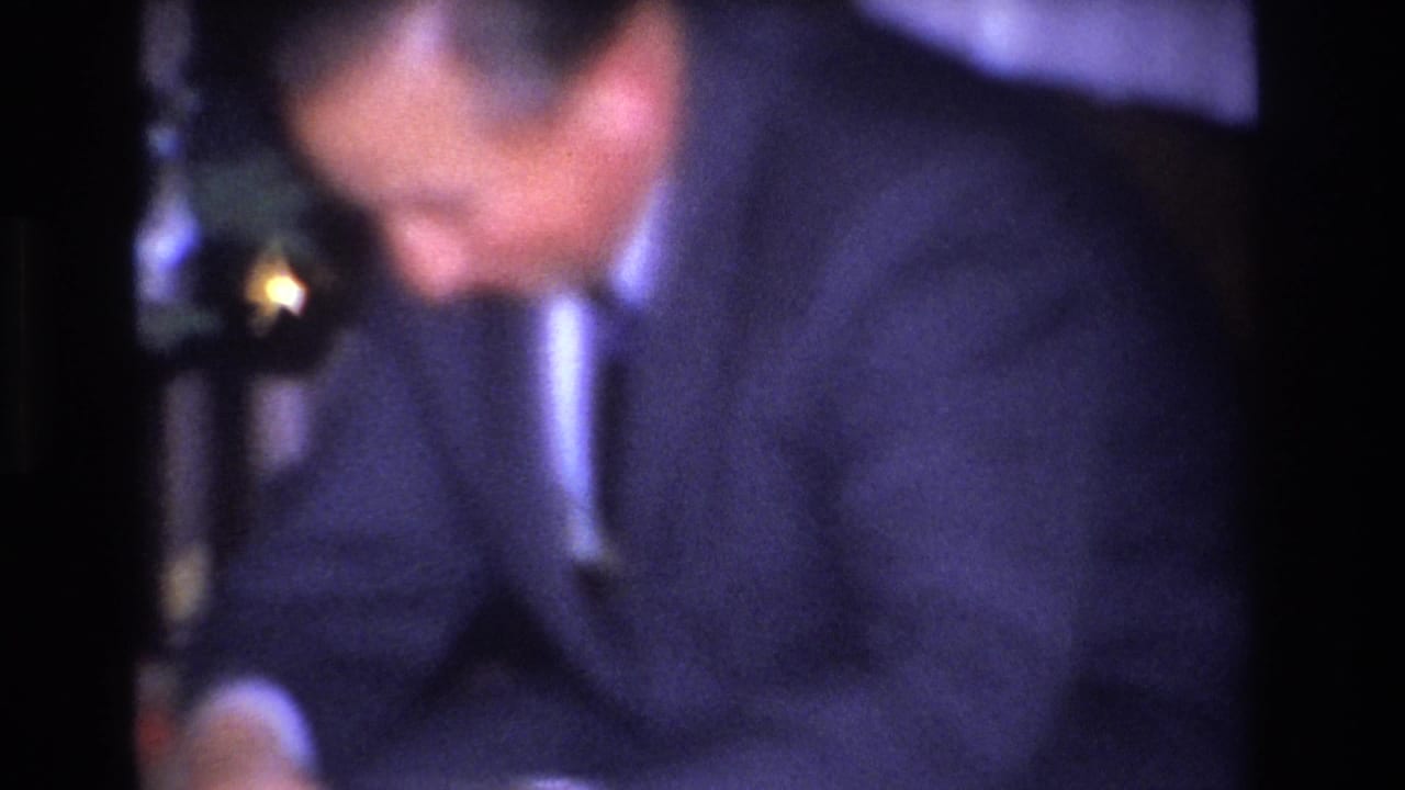 Man in dark suit and tie looking downward, Christmas tree branch with a gold ornament visible at upper right.