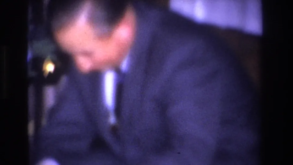 A formally dressed man in a dark suit and narrow tie looks downward in a mid-1960s indoor Christmas setting, 8mm home movie.