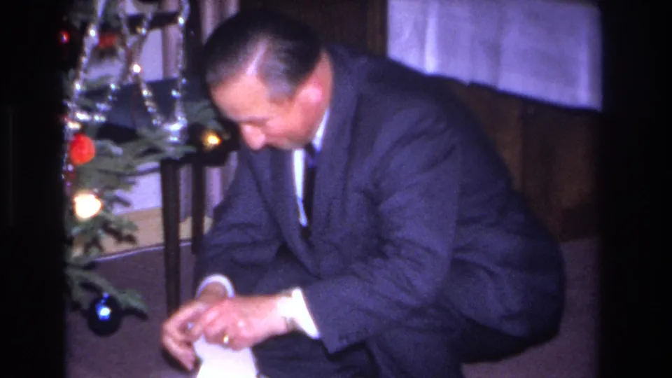 A formally dressed man in a dark suit and tie unwraps a package beside a Christmas tree with red and blue ornaments, in a private home interior, consistent with 1966 8mm footage.