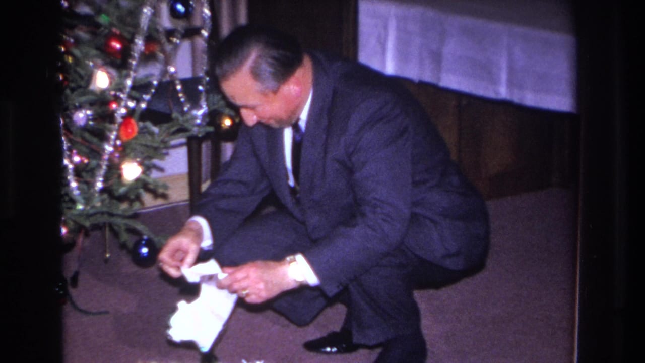 Man in dark suit opens a white-wrapped gift while kneeling on maroon carpet beside a tinsel-and-ornament Christmas tree; 8mm home movie, Hoboken, New Jersey, 1966.