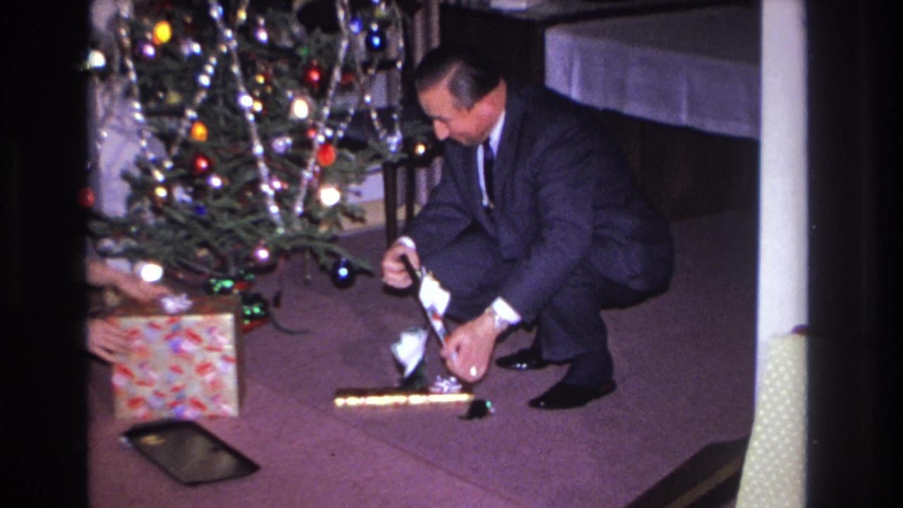Man in dark suit crouches near a decorated Christmas tree beside a colorful gift box and a dark flat rectangular tray on maroon carpet.