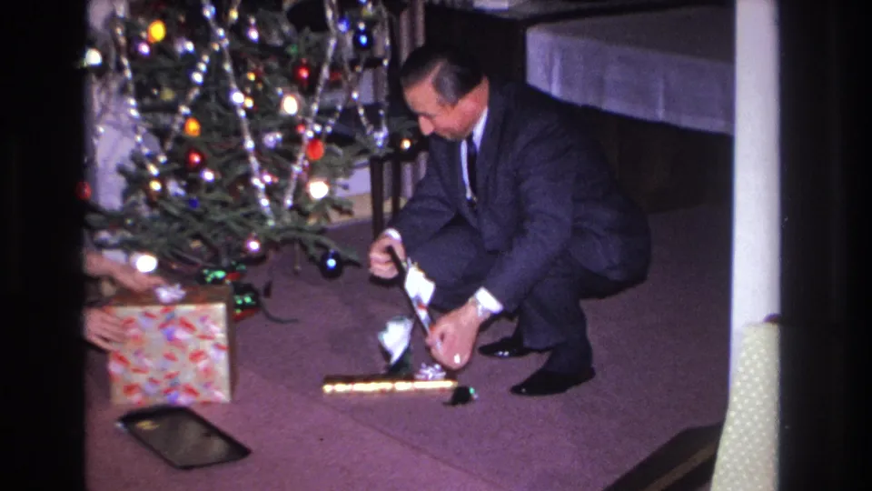 Man in dark suit handles wrapping paper beside a tinsel-covered Christmas tree; colorful patterned gift box and dark flat rectangular tray visible on maroon carpet; 8mm home movie, 1966.