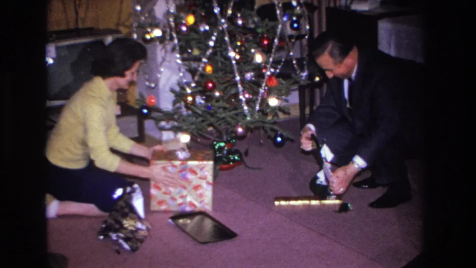 Man in dark suit and woman in yellow sweater open gifts together before a tinsel-decorated Christmas tree; period furniture visible at left; 8mm home movie, Hoboken, New Jersey, 1966.