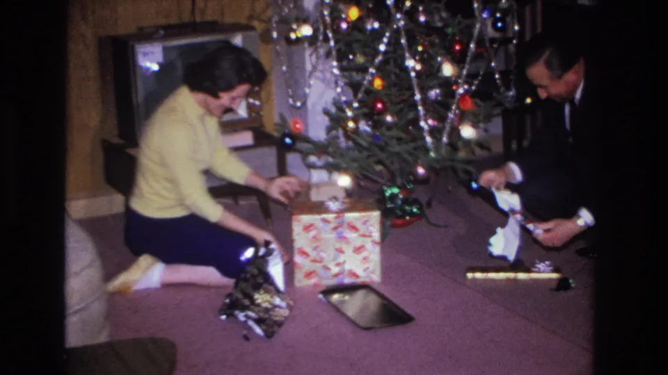 Woman in yellow sweater opens a gift beside a man in dark suit; decorated Christmas tree with colored lights and tinsel behind them; period furniture or TV visible at right; 8mm home movie, 1966.
