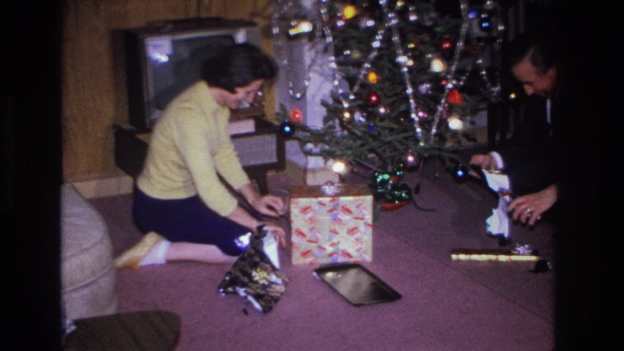 Woman in yellow sweater and man in dark suit kneeling by a lit Christmas tree with multicolored bulbs and silver tinsel, unwrapping gifts; 1966 Hoboken, New Jersey 8mm home movie.