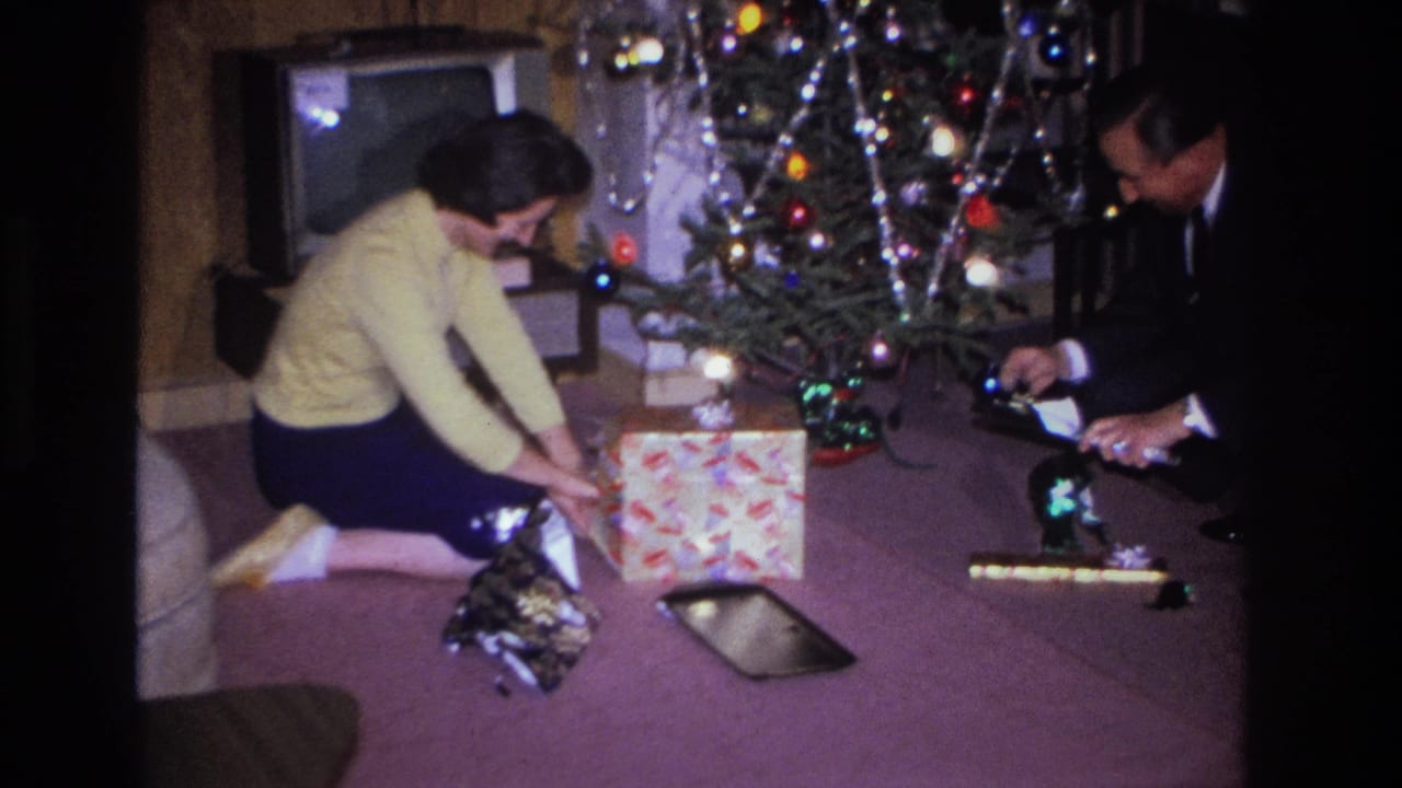 Woman in yellow sweater leaning over a wrapped gift near Christmas tree; man in dark suit partially visible at right.