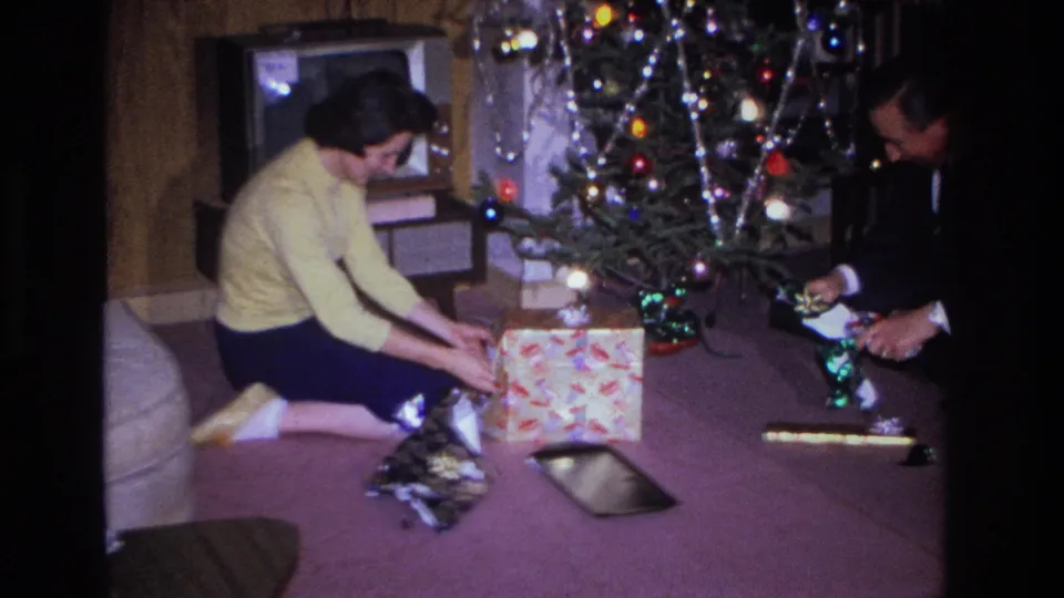 Woman in yellow sweater reaching toward festive gift boxes on purple carpet, man in dark suit at right; mid-1960s domestic Christmas interior, 8mm home movie.