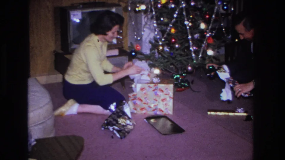 Woman in yellow sweater and man in dark suit beside a decorated Christmas tree with multicolored bulbs, unwrapping gifts on purple carpet; Hoboken, New Jersey, circa 1966.