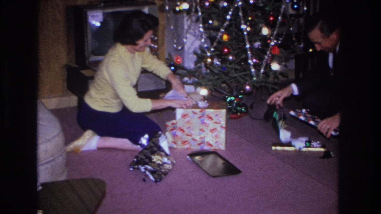 Couple opening Christmas gifts on the floor of a 1966 home interior in Hoboken, New Jersey, with a multicolored-lit tree and scattered wrapping paper behind them.