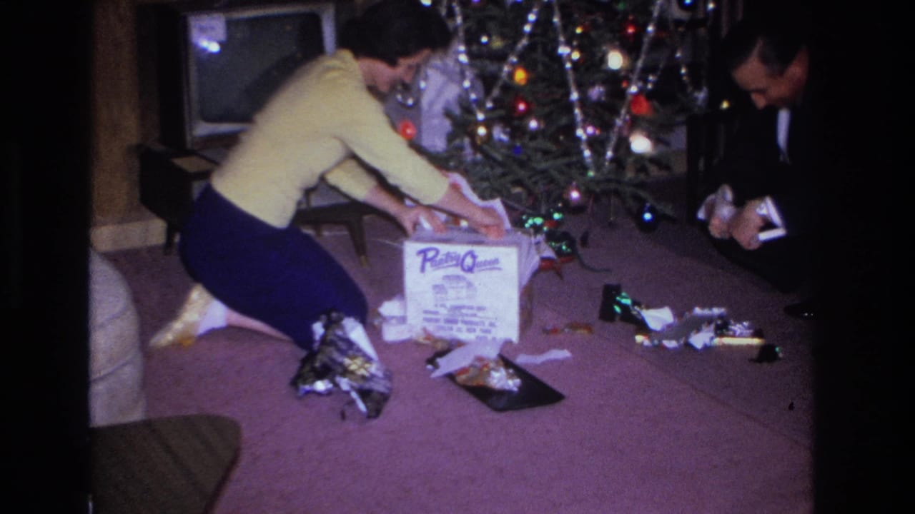 Woman reaches toward a white branded gift box among unwrapped presents beside a lit Christmas tree; man in dark suit visible at right.