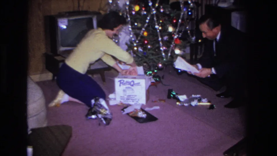 Couple kneeling on purple carpet opening gifts beside a lit 1960s Christmas tree; branded appliance box prominent among scattered wrapping, Hoboken, New Jersey, 1966.