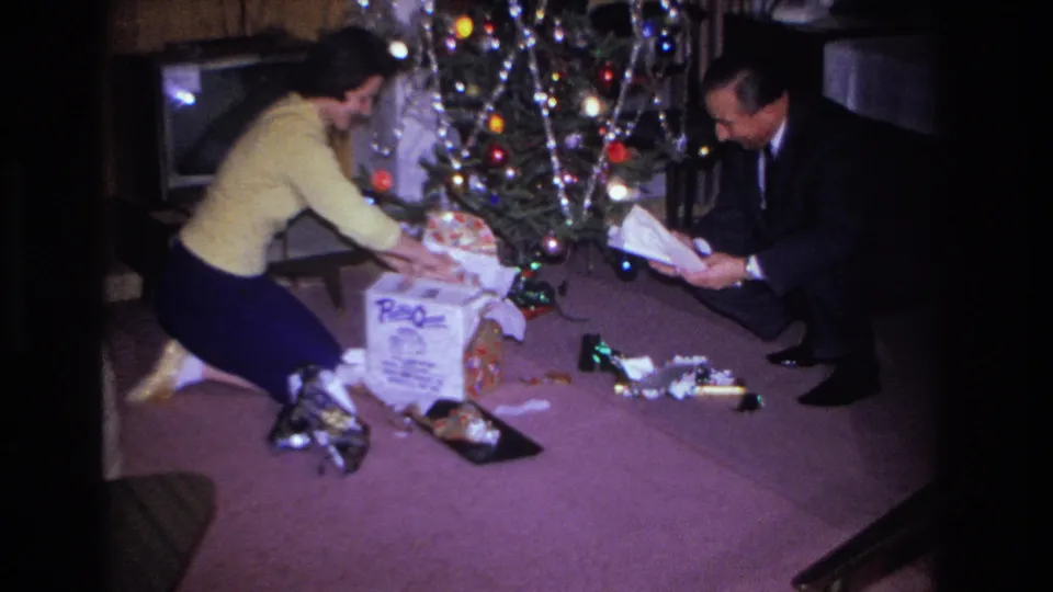 Woman in yellow sweater leaning forward among unwrapped gifts beside a 1960s Christmas tree; man in dark suit watching, branded box and console television visible, Hoboken, New Jersey, 1966.