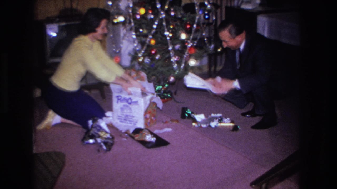 1966 home movie: a couple opens Christmas presents beside a tinsel-draped tree with colored lights in a mid-century American living room.