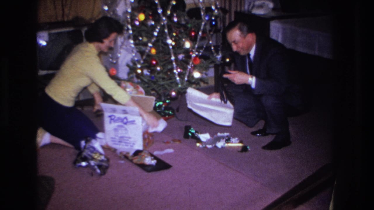 Couple at the Christmas tree; man in dark suit leans forward examining a gift while woman in yellow sweater reaches toward the packages.