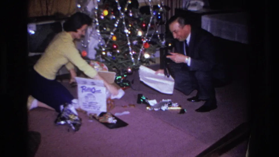 1966 8mm home movie: couple opening gifts beside a tinsel-draped Christmas tree with colored bulb lights in a private living room.