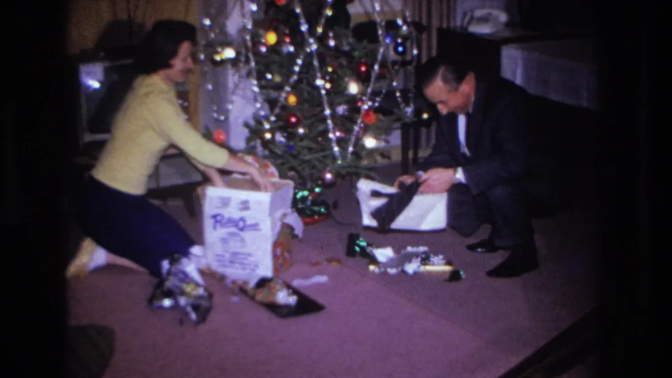 1966 home movie, closer view: couple engaged in Christmas gift opening beside a tinsel-decorated tree in a mid-century living room.