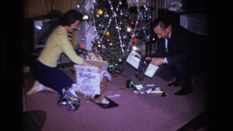 1966 8mm home movie: couple near a tinsel-draped Christmas tree with multicolored lights, surrounded by opened gifts and wrapping paper.