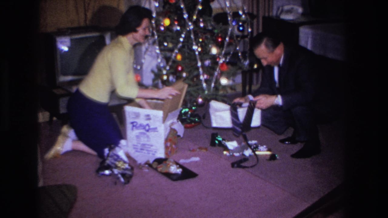 Woman and man on carpet beside a decorated Christmas tree examining a large white gift box; television set visible in background.