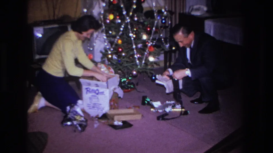 Adult couple bent over a large printed gift box on a purple carpet in a 1960s living room; Christmas tree with multicolored bulb lights and foil tinsel in the background, circa 1966.
