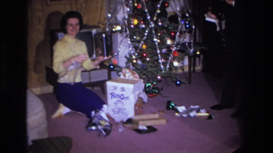 Woman in yellow sweater smiling toward camera while seated on a purple carpet beside a decorated Christmas tree in a private home interior, circa 1966; a third person's lower body is visible at the right edge.